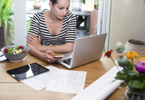 Frau am Schreibtisch mit Laptop und Mobiltelefon neben einem Bauplan und Salat - REAF000113
