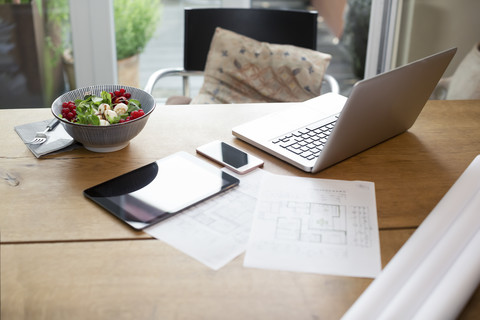 Schreibtisch mit Laptop und Mobiltelefon neben Bauplan und Salat, lizenzfreies Stockfoto