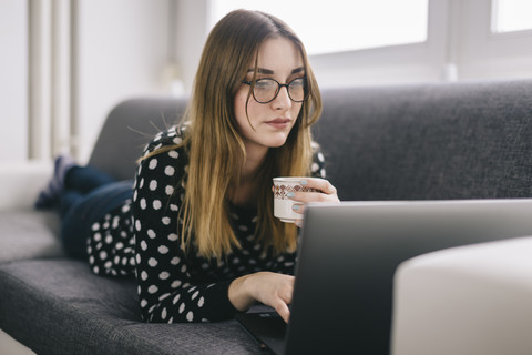 Young woman relaxing on the couch with cup of coffee using laptop stock photo