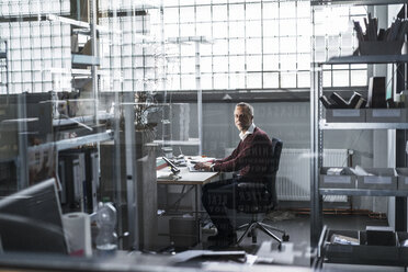 Älterer Mann mit Laptop am Schreibtisch in einer Fabrik - KNSF000148