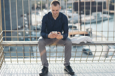 Businessman sitting on bench in front of harbour text messaging - SKCF000141