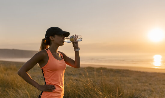 Junge Sportlerin, die bei Sonnenuntergang Wasser trinkt - MGOF002139