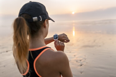 Junge Sportlerin, die am Strand bei Sonnenuntergang auf ihre Smartwatch schaut - MGOF002155