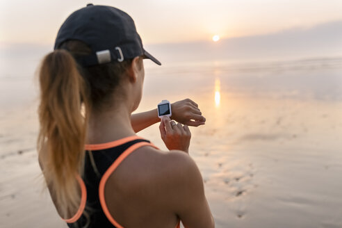 Young athlete woman looking the smartwatch on the beach at sunset - MGOF002155