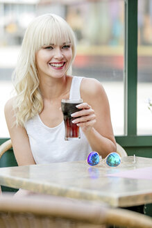 Porträt einer lächelnden blonden Frau, die in einem Straßencafé sitzt und Cola trinkt - GDF001103