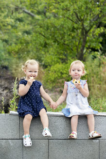 Portrait of two little sisters sitting hand in hand on a wall eating pastries - HAPF000683