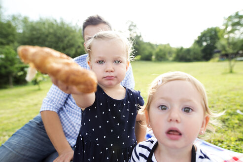 Little girl with father and sister in a park offering croissant to viewer - HAPF000706