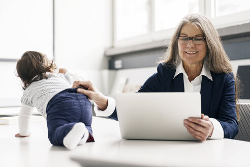 Senior businessswoman sitting at conference table with laptop and baby girl - KNSF000183