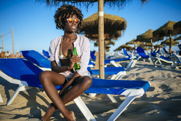 Junge Frau sitzt am Strand und trinkt Bier - KIJF000670