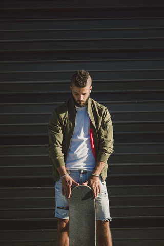 Mann mit Skateboard steht vor einer Fassade und schaut nach unten, lizenzfreies Stockfoto