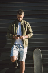 Mann mit Skateboard steht vor einer Fassade und schaut auf sein Handy - RAEF001342