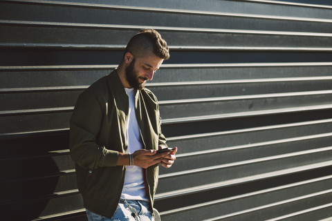 Lächelnder Mann, der vor einer Fassade steht und auf sein Mobiltelefon schaut, lizenzfreies Stockfoto