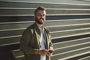 Portrait of smiling man with cell phone standing in front of facade - RAEF001344