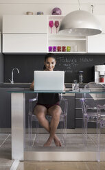 Young woman sitting at table in the kitchen working with laptop - MOMF000017