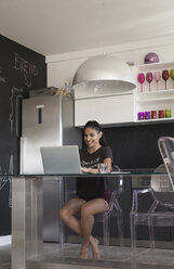 Young woman sitting at table in the kitchen working with laptop - MOMF000018