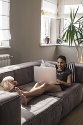 Junge Frau sitzt mit ihrem Hund auf der Couch und benutzt einen Laptop - MOMF000025