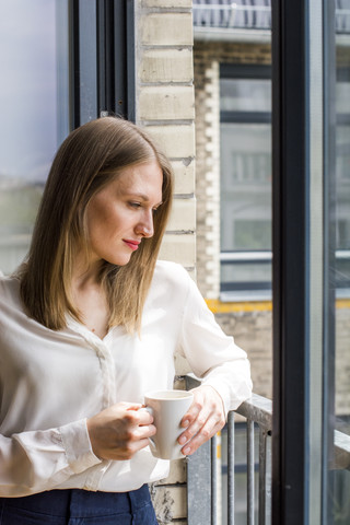 Frau mit Kaffeetasse steht am offenen Fenster und schaut nach unten, lizenzfreies Stockfoto