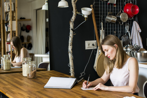 Frau sitzt am Tisch in ihrer Küche und schreibt etwas auf, lizenzfreies Stockfoto