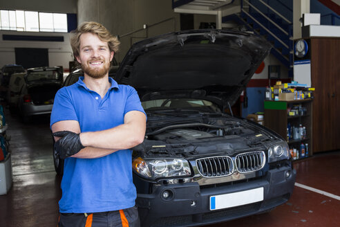 Selbstbewusster Mechaniker, der mit verschränkten Armen in seiner Autowerkstatt steht - ABZF000967