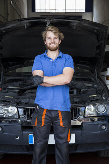 Selbstbewusster Mechaniker, der mit verschränkten Armen in seiner Autowerkstatt steht - ABZF000971