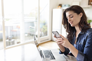 Smiling young woman at home using cell phone and laptop - PESF000275