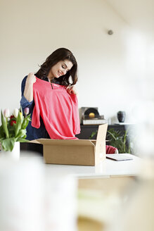 Lächelnde Frau zu Hause beim Auspacken eines Pakets mit Kleidungsstücken - PESF000343