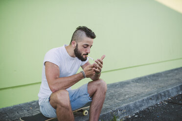 Junger Mann sitzt auf einem Skateboard und zündet sich eine Zigarette an - RAEF001372