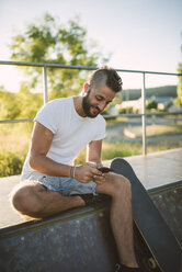 Lächelnder Skateboarder benutzt sein Handy in einem Skatepark - RAEF001375