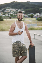 Portrait of smiling man with skateboard aand smartphone - RAEF001394