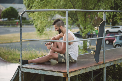 Lächelnder Skateboarder benutzt sein Smartphone in einem Skatepark, lizenzfreies Stockfoto