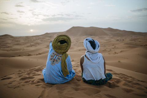 Frau sitzt mit Berberführer in der Wüste und beobachtet den Sonnenuntergang, lizenzfreies Stockfoto