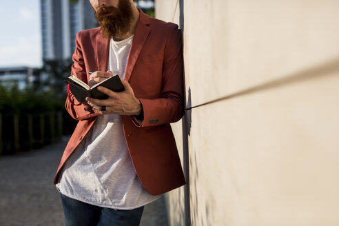 Modischer junger Mann mit Vollbart lehnt an der Wand und schreibt in sein Notizbuch - MAUF000729