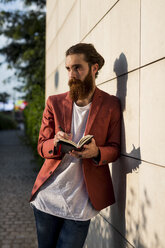 Modischer junger Mann mit Notebook, der an der Wand lehnt und in die Ferne schaut - MAUF000730