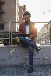 Junger Mann sitzt auf einer Mauer in der Stadt und benutzt ein Tablet - MAUF000741