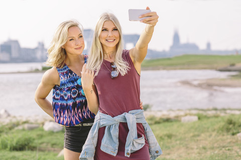 Zwei lächelnde junge Frauen machen ein Selfie am Flussufer, lizenzfreies Stockfoto