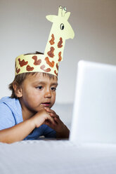 Kleiner Junge mit selbstgebasteltem Kopfschmuck liegt auf dem Bett und schaut auf den Laptop - VABF000744