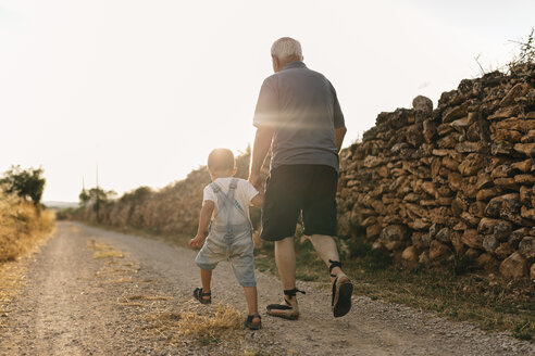 Back view of little boy and his great-grandfather walking on dirt track at backlight - JRFF000816