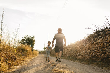 Kleiner Junge und sein Urgroßvater gehen auf einem Feldweg bei Gegenlicht - JRFF000817