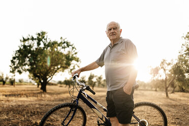 Senior man with bicycle standing on a field at evening twilight - JRFF000819