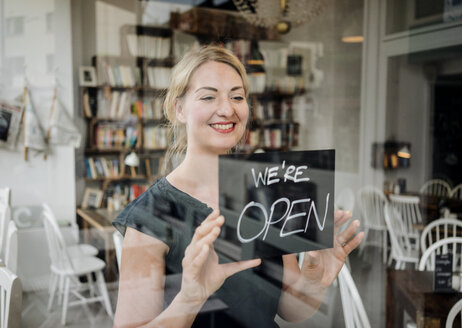 Lächelnde Frau in einem Café, die ein offenes Schild an einer Glasscheibe befestigt - KNSF000204