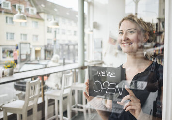 Lächelnde Frau in einem Café, die ein offenes Schild an einer Glasscheibe befestigt - KNSF000205