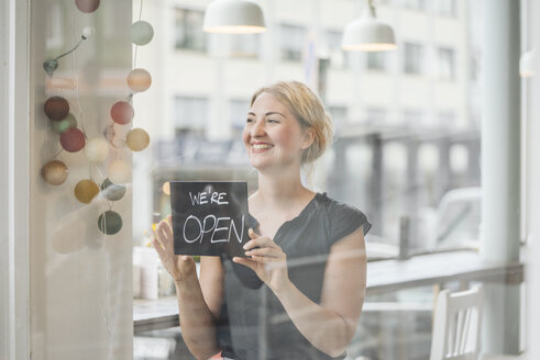 Lächelnde Frau in einem Café, die ein offenes Schild an einer Glasscheibe befestigt - KNSF000233
