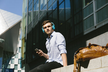 Ireland, Dublin, young businessman sitting on a wall listening music with headphones - BOYF000513