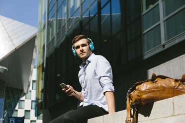 Irland, Dublin, junger Geschäftsmann, der auf einer Mauer sitzt und mit Kopfhörern Musik hört - BOYF000513