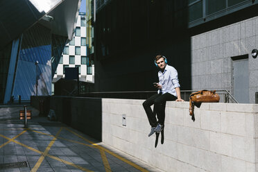 Ireland, Dublin, young businessman sitting on a wall listening music with headphones - BOYF000514