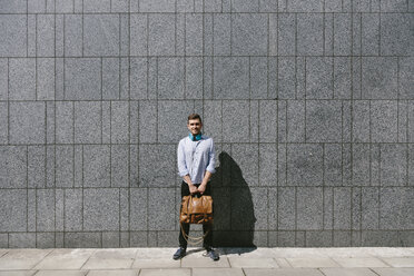 Smiling young businessman standing in front of a grey wall holding leather bag - BOYF000515