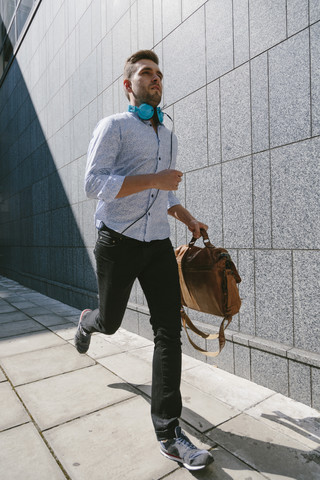 Laufender junger Geschäftsmann mit Kopfhörern und Ledertasche, lizenzfreies Stockfoto