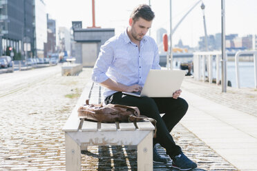 Irland, Dublin, junger Geschäftsmann sitzt auf einer Bank und benutzt einen Laptop - BOYF000524