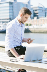 Irland, Dublin, junger Geschäftsmann sitzt auf einer Bank und benutzt Laptop und Handy - BOYF000528