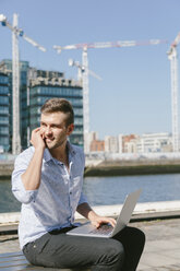 Ireland, Dublin, smiling young businessman sitting on bench with laptop telephoning with cell phone - BOYF000529